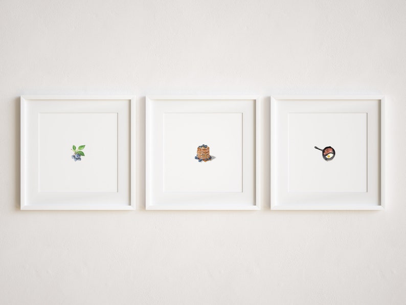 May include: Three white picture frames with white mats hanging on a white wall. The left frame shows a small sprig of blueberries. The middle frame shows a stack of pancakes with blueberries on top. The right frame shows a frying pan with a fried egg inside.