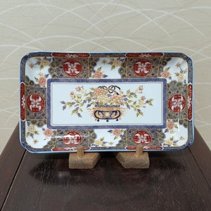 May include: A rectangular ceramic serving tray with a white background and a colorful floral design. The tray has a gold trim and a blue and red border.