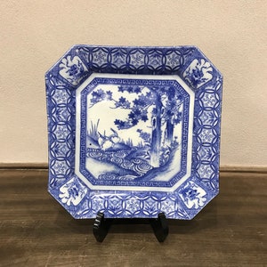 May include: A blue and white porcelain plate with an octagonal shape. The plate features a Japanese-inspired design with a tree, water, and birds. The border of the plate is decorated with a repeating pattern of circles and squares.