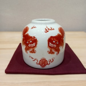 Puede incluir: Jarrón de cerámica blanca con un diseño rojo y blanco de dos leones y llamas. El jarrón está sentado sobre un paño rojo.