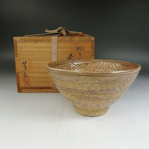 May include: A tan ceramic bowl with a textured, diagonal pattern. The bowl sits in front of a wooden box with Japanese characters and a ribbon. The bowl has a wide opening and a rounded base.