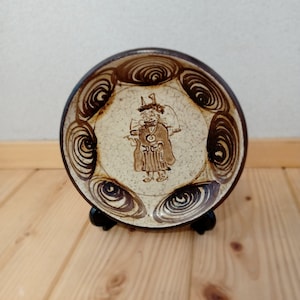 May include: A small, round, white ceramic plate with a brown and white design. The plate features a stylized figure in the center and a swirling pattern around the edge. The plate is on a wooden stand.
