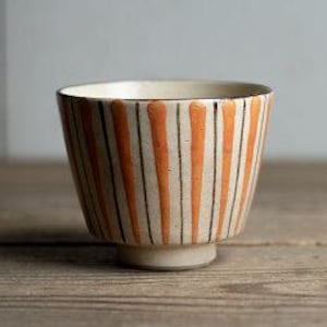 May include: A small, white ceramic bowl with orange and black vertical stripes. The bowl has a rounded bottom and a slightly flared rim.
