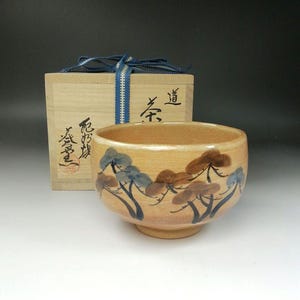 Könnte beinhalten: Eine beige Keramik-Teeschale mit handgemaltem Baumdesign in Blau und Braun. Die Schale steht vor einer Holzkiste, die mit einem blauen Band gebunden ist. Die Box hat japanische Schriftzeichen.
