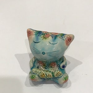 以下が含まれることがあります： 目を閉じた青い猫の陶器の置物で、足に魚を持っています。猫はカラフルな花の模様で飾られています。