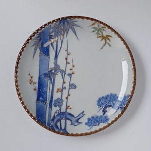 以下が含まれることがあります： 茶色の縁取りと青と金の色の花柄が施された白い陶磁器の皿。デザインは竹の茎、松の木、花を特徴としています。