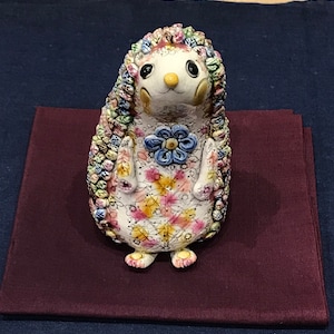 Könnte beinhalten: Eine Keramikfigur eines Igels mit weißem Körper und einem bunten Blumenmuster. Der Igel hat eine Blume auf der Brust und einen verspielten Ausdruck.