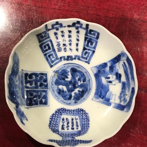 May include: A white porcelain bowl with a blue and white design. The bowl has a scalloped edge and features a central floral motif surrounded by four smaller motifs. The motifs include a dragon, a building, a landscape, and a geometric pattern. The bowl is decorated with Chinese characters.