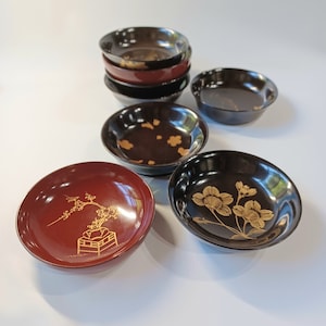 以下が含まれることがあります： 金色の花柄が施された、黒と赤の漆塗りの小さな丸いボウル6個セット。ボウルはピラミッド型に積み重ねられており、一番下のボウルが一番大きく、一番上のボウルが一番小さい。