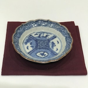 May include: A blue and white ceramic plate with a scalloped edge and a brown rim. The plate features a detailed design with birds and geometric patterns. It rests on a dark red cloth, creating a contrast in color and texture.