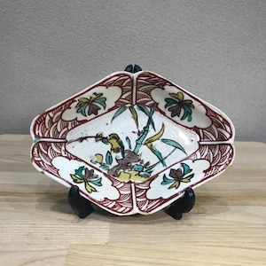 Könnte beinhalten: Rautenformige Keramikschale mit bemaltem Design. Der Rand hat ein rot-weißes Muster. Die Mitte zeigt einen Vogel, Bambus und florale Elemente in Grün, Gelb und Braun. Die Schale steht auf einem schwarzen Ständer.
