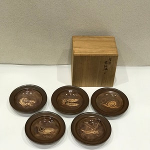 May include: Set of five small brown ceramic plates with a raised rim. Each plate features a different insect design in a light brown color. The plates are arranged in a circle on a white surface. A wooden box with Japanese writing is in the background.
