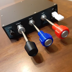 May include: A black control panel with silver, black, blue, and red knobs and levers. The panel has labels including "TRIM", "THROT", "PROP", and "MIX". The levers are for controlling various functions. The panel is on a wooden surface.