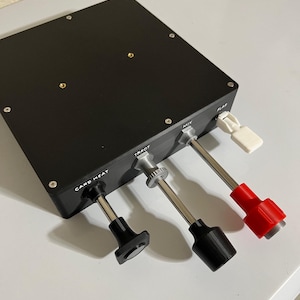 May include: A black control panel with silver and black levers labeled "CARD HEAT", "THROT", "MIX", and "FLAP". The "FLAP" lever has a white handle, and the "CARD HEAT" lever has a black handle. The panel is on a white surface.