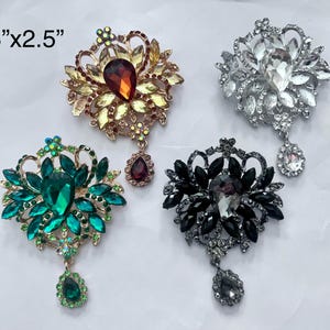 May include: Four decorative brooches with jeweled accents. The brooches are in the shapes of flowers with a single large jewel in the center of each. The jewels are green, red, black, and clear. The brooches are 3 inches by 2.5 inches.