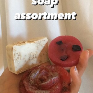 May include: Assortment of three handmade soaps. One soap is a light brown and white bar with a textured surface. Another soap is a red rose shape with a glittery finish. The third soap is a round, red soap with black flecks.