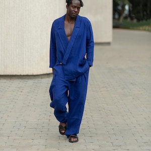 Może przedstawiać: Osoba w żywym, niebieskim lnianym garniturze, składającym się z marynarki i pasujących spodni. Marynarka ma unikalne zapięcie na zakładkę. Strój uzupełniają czarne sandały. Tło jest zewnętrzne, z neutralnym tłem.