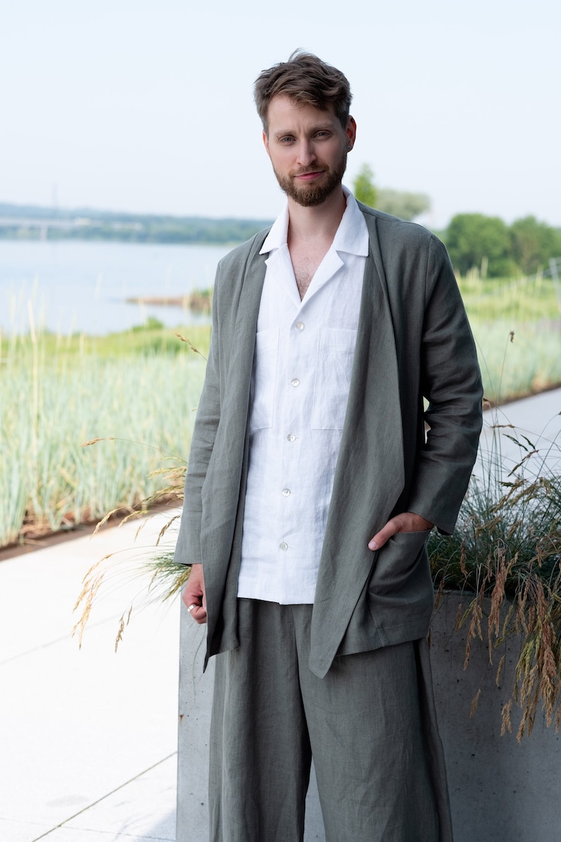 Peut inclure: Un homme portant un costume en lin gris clair avec une chemise en lin blanche. La veste de costume est ouverte et l'homme est debout, les mains dans les poches.