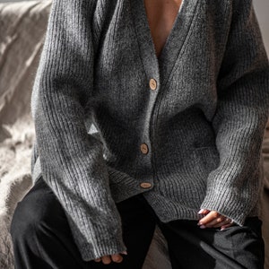 K&ouml;nnte beinhalten: Grauer Strickcardigan mit Rippenmuster, V-Ausschnitt und Holzkn&ouml;pfen. Der Pullover hat eine lockere Passform und eine Tasche auf der Vorderseite. Die Person tr&auml;gt eine schwarze Hose.
