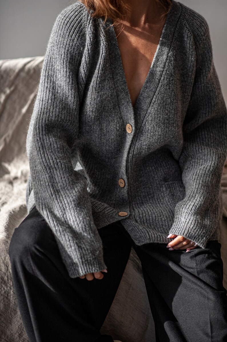 K&ouml;nnte beinhalten: Grauer Strickcardigan mit Rippenmuster, V-Ausschnitt und Holzkn&ouml;pfen. Der Pullover hat eine lockere Passform und eine Tasche auf der Vorderseite. Die Person tr&auml;gt eine schwarze Hose.