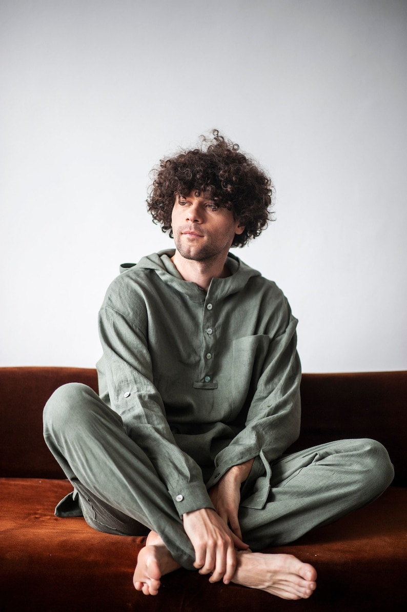 May include: A person wearing a green hooded linen jumpsuit, sitting on a brown couch. The person has curly brown hair and is looking to the side.