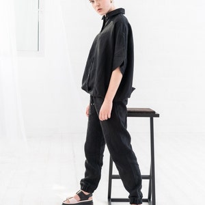 K&ouml;nnte beinhalten: Eine Frau tr&auml;gt ein schwarzes Leinenhemd und eine passende Hose. Sie steht auf einem schwarzen Metallhocker und tr&auml;gt schwarze Sandalen mit einem rosafarbenen Riemen.