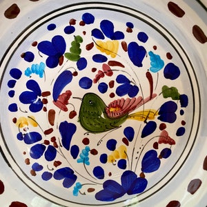 Pode incluir: Uma tigela de cer&acirc;mica branca com uma borda azul e marrom. A tigela &eacute; decorada com um desenho pintado &agrave; m&atilde;o de um p&aacute;ssaro verde com detalhes amarelos e vermelhos, rodeado de flores azuis, vermelhas, amarelas e verdes.