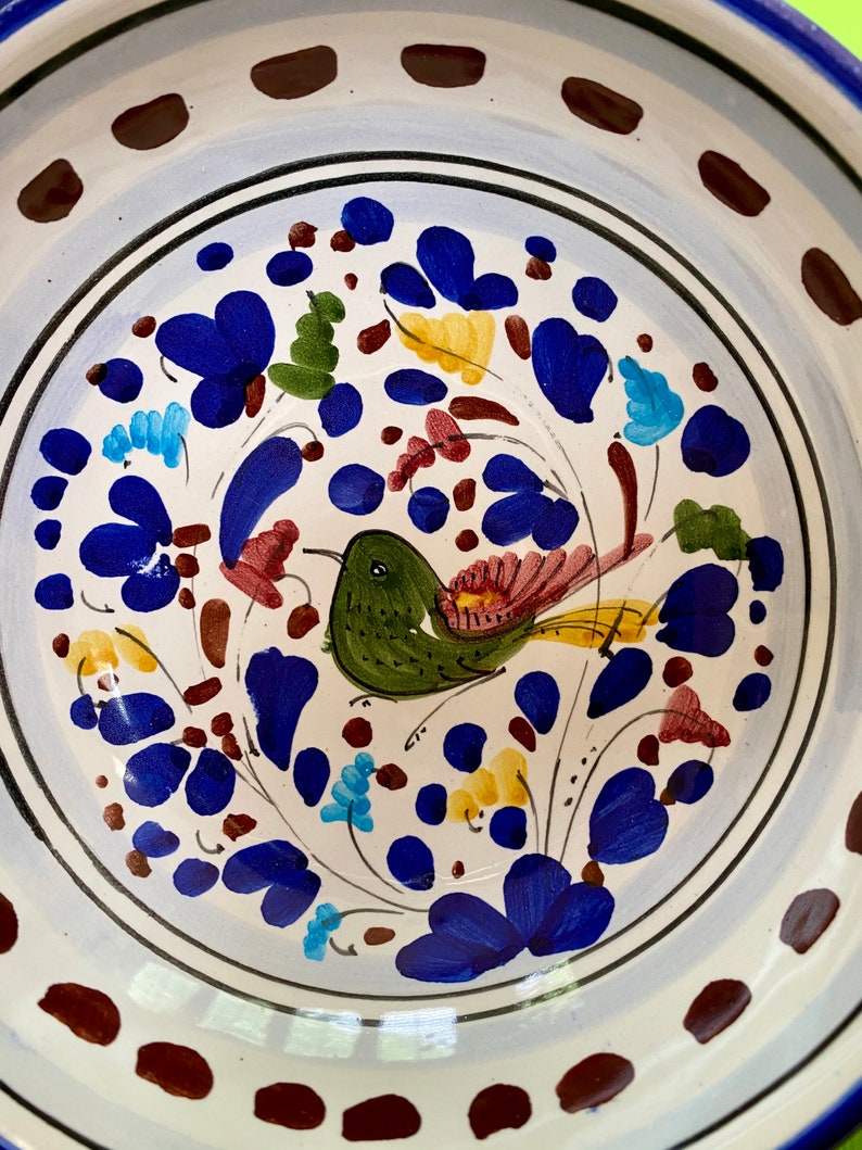 Pode incluir: Uma tigela de cer&acirc;mica branca com uma borda azul e marrom. A tigela &eacute; decorada com um desenho pintado &agrave; m&atilde;o de um p&aacute;ssaro verde com detalhes amarelos e vermelhos, rodeado de flores azuis, vermelhas, amarelas e verdes.