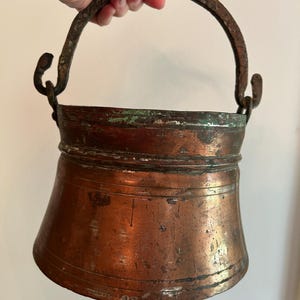 May include: A copper-colored metal bucket with a curved handle. The bucket has a flared shape and shows signs of age and wear. The handle is made of a darker, textured metal.