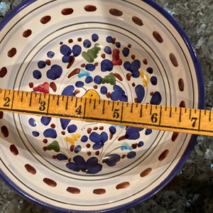Pode incluir: Uma tigela de cer&acirc;mica branca com uma borda azul e um desenho floral em tons de azul, vermelho, amarelo e verde. A tigela &eacute; decorada com pontos marrons ao redor da borda e tem um di&acirc;metro de aproximadamente 18 cm.