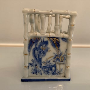 May include: A small white ceramic vase with a blue and gold bamboo design. The vase is shaped like a small fence with a decorative design on the front.