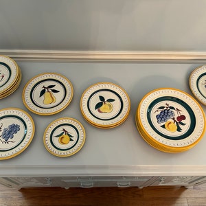 May include: A set of nine vintage ceramic plates with yellow rims and green and blue fruit designs. The plates feature pears, grapes, cherries, and peaches.