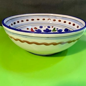 Pode incluir: Uma tigela de cer&acirc;mica branca com uma borda azul e marrom e um desenho floral em azul, vermelho e verde. A tigela est&aacute; sobre uma superf&iacute;cie verde.