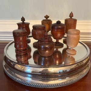 May include: A collection of small, handcrafted wooden jars with lids, displayed on a silver-rimmed mirrored tray. The jars vary in shape and wood grain, showcasing a range of natural tones from light to dark brown. The tray sits on a dark wooden surface.