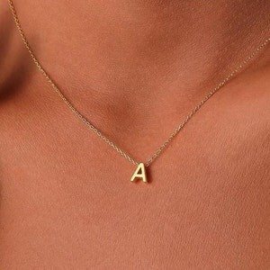 May include: A delicate gold-colored necklace featuring the letter "A" pendant. The pendant is small and centered on a fine chain. The necklace is worn around the neck, with the chain resting against the skin.