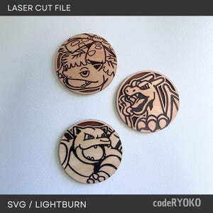 Peut inclure: Trois cercles en bois découpés au laser représentant des personnages Pokémon. Le cercle supérieur montre Bulbizarre, à droite Dracaufeu et en bas Tortank. Le texte "LASER CUT FILE" et "SVG / LIGHTBURN" sont visibles.