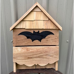 May include: A wooden bat house with a black bat silhouette on the front. The house has a shingled roof and a small opening for bats to enter.