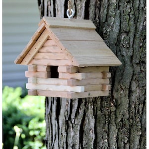 May include: A wooden birdhouse shaped like a log cabin with a small opening for birds to enter. The birdhouse is hanging from a tree branch.
