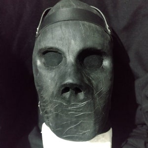 Puede incluir: Una máscara negra con una superficie texturizada y una correa negra. La máscara tiene agujeros para los ojos y un agujero para la boca.