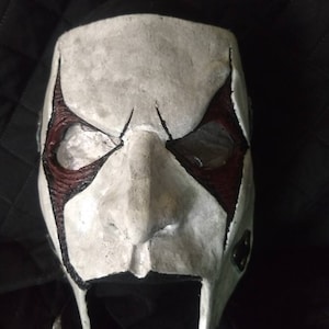 May include: A full-face mask featuring a textured white base with prominent black and dark red accents around the eye sockets. The mask has a sculpted nose, defined brow lines, and two downward-pointing extensions at the chin area.