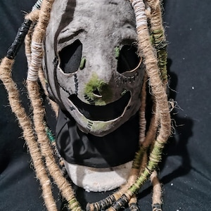 May include: A gray mask with stitched details and large eye openings. The mask has a smiling mouth and is adorned with long, rope-like dreadlocks in shades of brown, green, and black. The mask appears to be handmade.