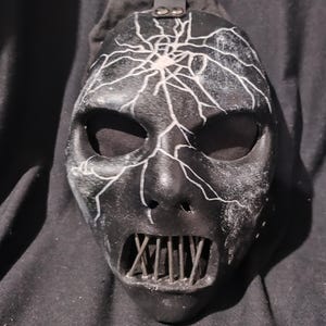 Puede incluir: Una máscara negra y blanca con un diseño agrietado y una protección bucal de metal. La máscara tiene un patrón de rayo blanco en la frente y una protección bucal de metal con múltiples barras.