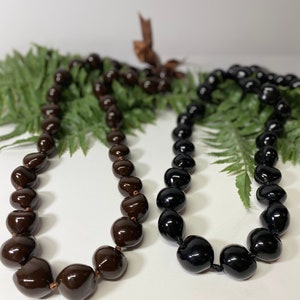 May include: Two necklaces made of large, glossy beads. One necklace is dark brown, the other is black. Both necklaces are strung on a dark cord. The necklaces are displayed on a white surface with green fern leaves.