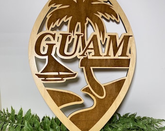 May include: Wooden wall art featuring the word "GUAM" in a cutout design. The artwork includes a palm tree, sailboat, and island scene, all in a natural wood tone against a lighter wood background. Decorative item.
