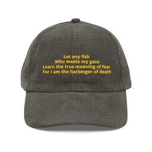 May include: A dark gray corduroy baseball cap featuring yellow embroidered text that reads: Let any fish Who meets my gaze Learn the true meaning of fear For I am the harbinger of death. The cap has a structured crown and a curved brim.