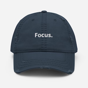 Może przedstawiać: Znoszona granatowa czapka z daszkiem z białym haftem "Focus". Czapka ma wygięty daszek i zużyty, vintage wygląd z postrzępionymi krawędziami.