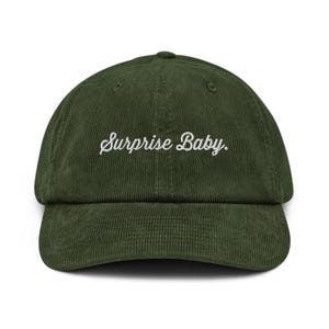 May include: A dark olive green corduroy baseball cap featuring the text "Surprise Baby." embroidered in white cursive script across the front center. The hat has a structured crown, a curved brim, and a classic six-panel design with eyelets.