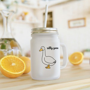 Puede incluir: Taza de vidrio blanco tipo mason con tapa plateada y pajita blanca. La taza presenta una ilustración de dibujos animados de un ganso con las palabras "silly goose" en negro. La taza tiene un asa y está sobre una superficie de madera.
