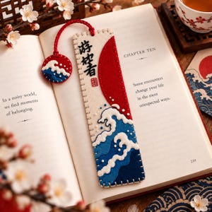 May include: A rectangular felt bookmark featuring an embroidered design of a large red sun and blue ocean waves. The bookmark includes a matching circular felt charm attached by a red cord. Black Japanese characters are stitched vertically.
