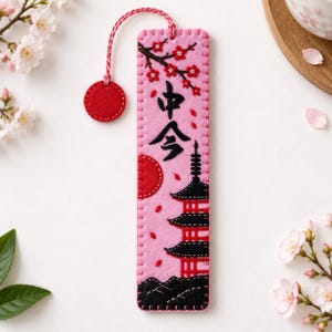 May include: A rectangular pink felt bookmark featuring black embroidered Japanese kanji characters, a red sun, a black and red pagoda, and a cherry blossom branch. The bookmark has a pink braided cord with a circular red felt tab at the top.